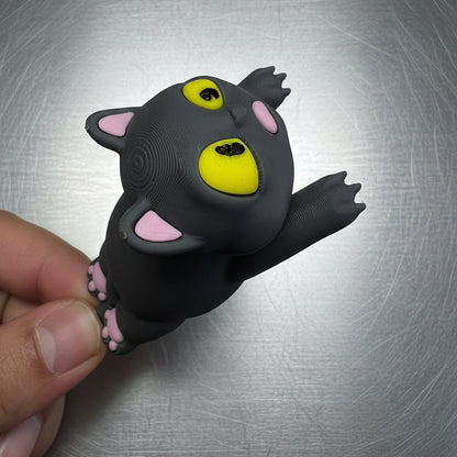 Black cat-shaped toy with yellow eyes and pink ears held by a hand on a metallic surface