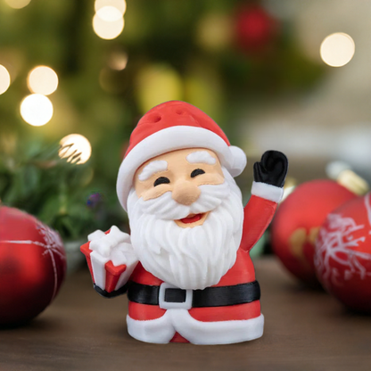 A 3D printed Santa Claus figure with a red outfit and white beard, smiling and waving, placed on a table with Christmas decorations in the background.